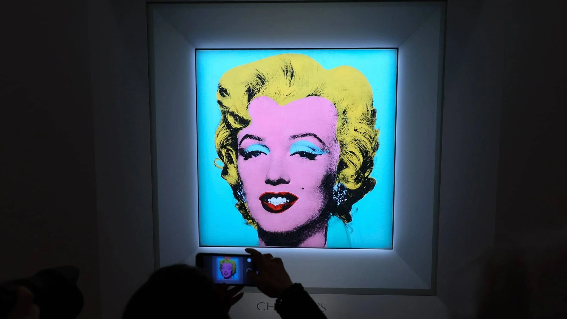 Visitante fotografía con su móvil la icónica serigrafía de Marilyn Monroe de Warhol, expuesta iluminada en sala oscura de Christie's.