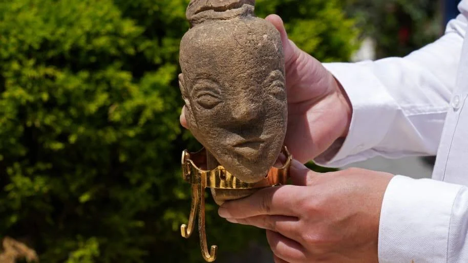Manos sostienen una antigua figura de piedra con rostro tallado y un adorno de metal dorado en la parte inferior.