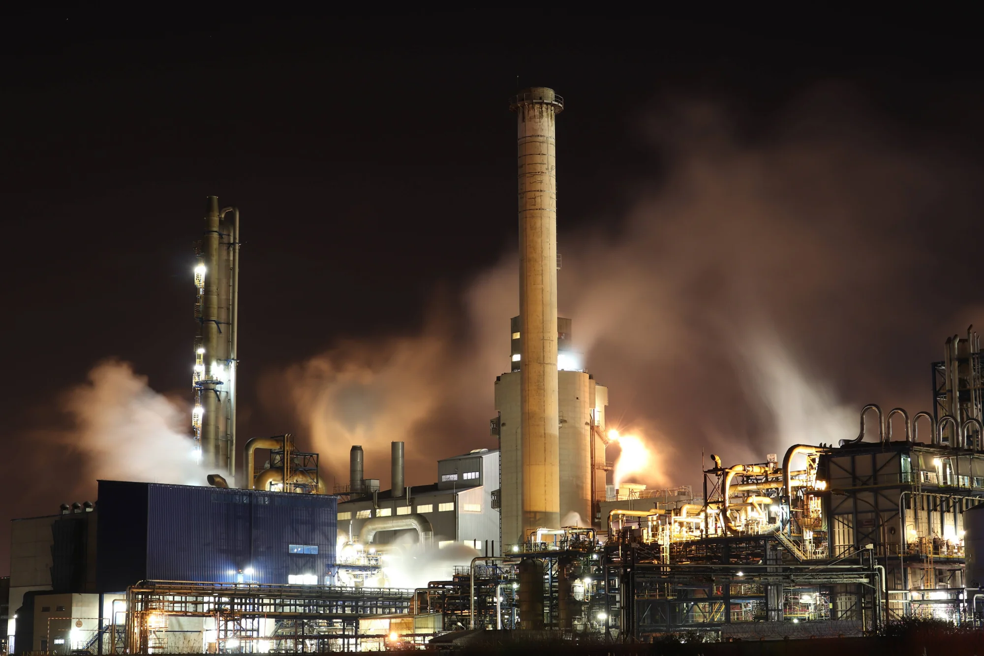 Complejo industrial iluminado de noche con chimeneas expulsando vapor y humo al cielo oscuro.