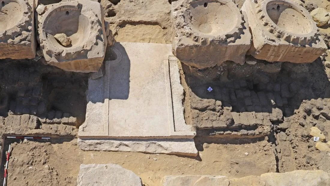 Vista aérea de una excavación arqueológica que muestra una losa de piedra clara rodeada de bloques de piedra tallados en terreno arenoso.