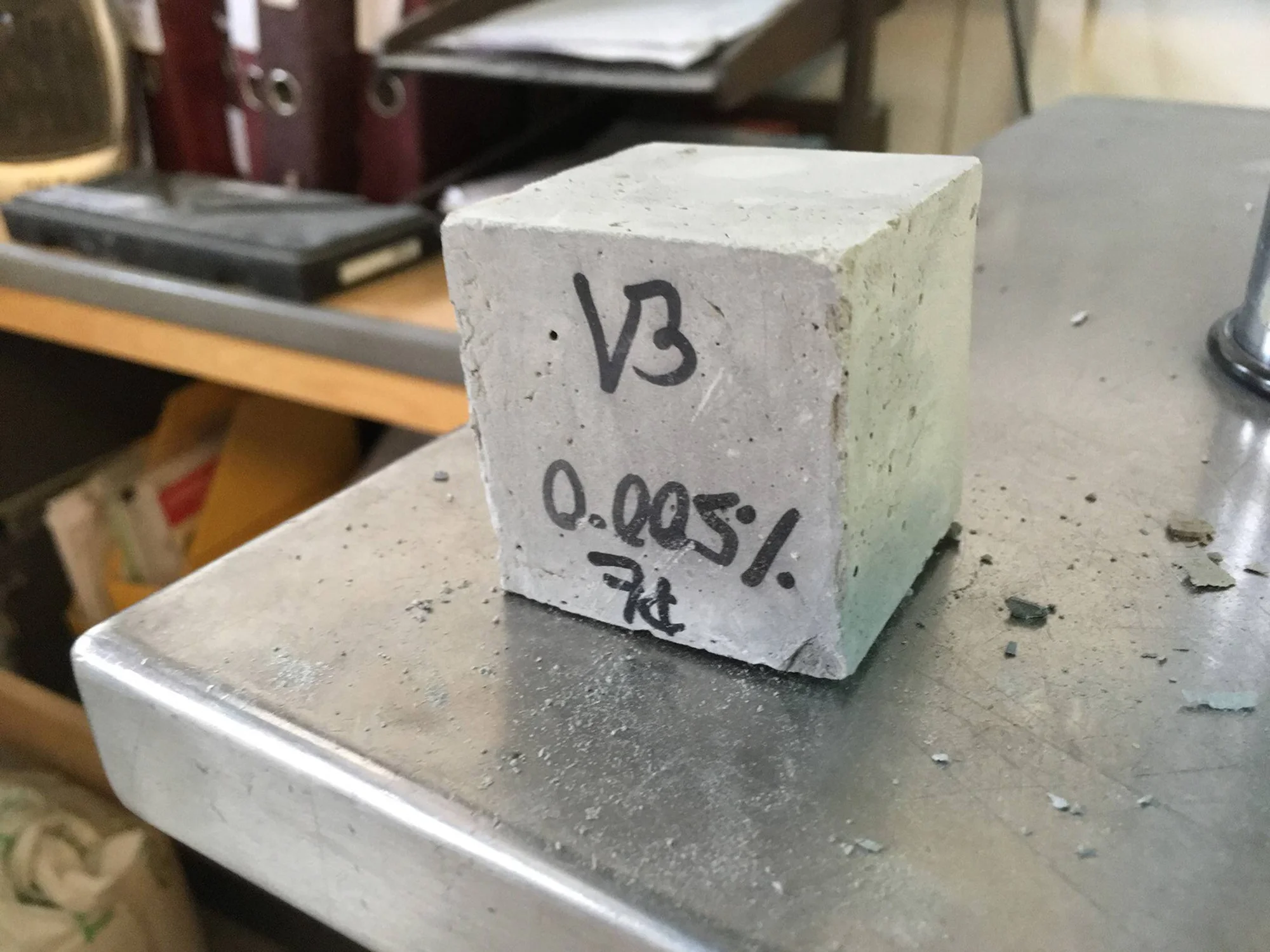 Cubo de hormigón con la inscripción 'V3 / 0.005% / 7kt' apoyado sobre una superficie metálica en un laboratorio.