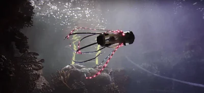 Robot submarino con estructura de brazos iluminados con LEDs rojos flotando sobre rocas en el fondo del mar.