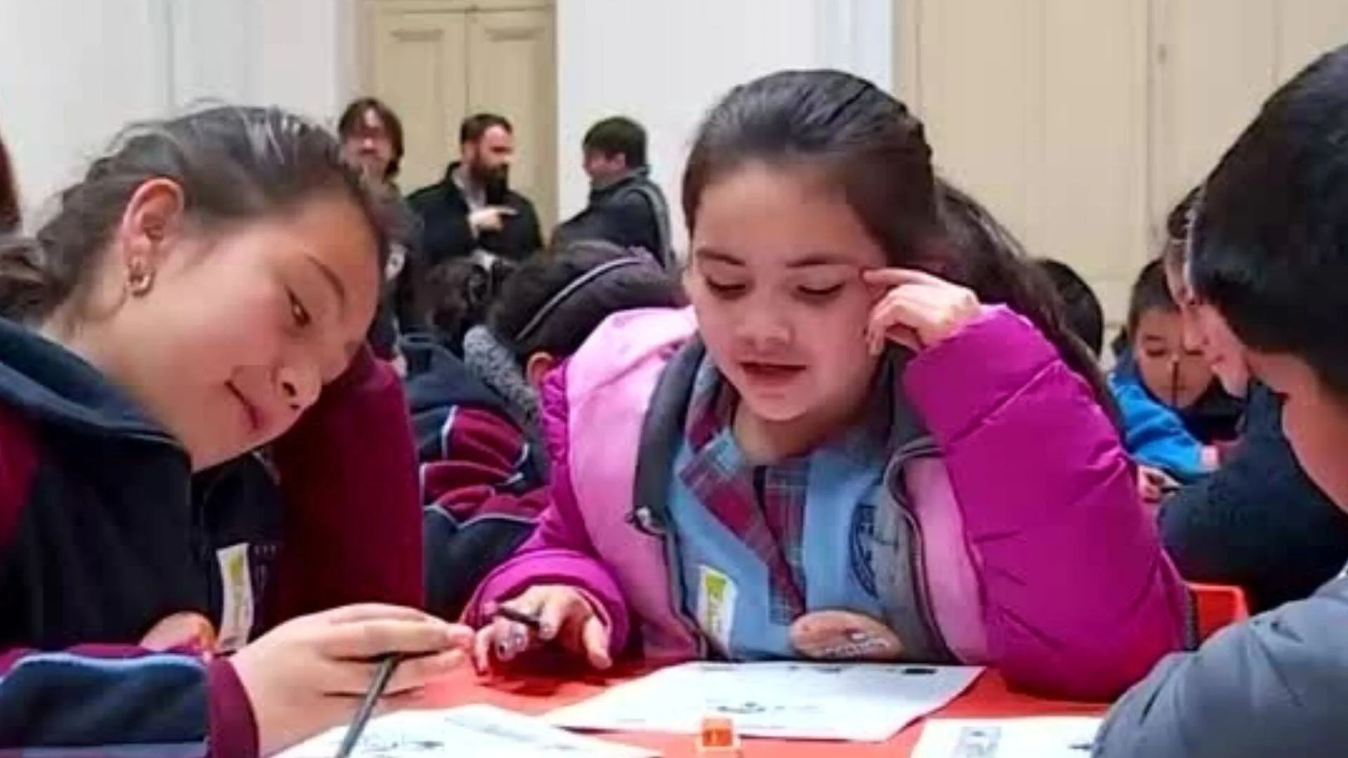Dos niñas concentradas resuelven una actividad en papel sobre una mesa roja, rodeadas de otros estudiantes.