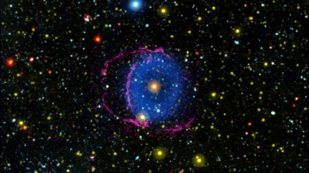 Nebulosa planetaria con anillo de gas magenta rodeando una nube azul y una estrella central naranja, captada en falso color.