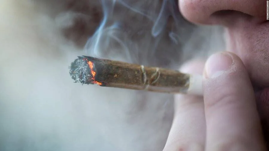 Primer plano de una persona sosteniendo y fumando un cigarrillo de marihuana encendido con humo visible.
