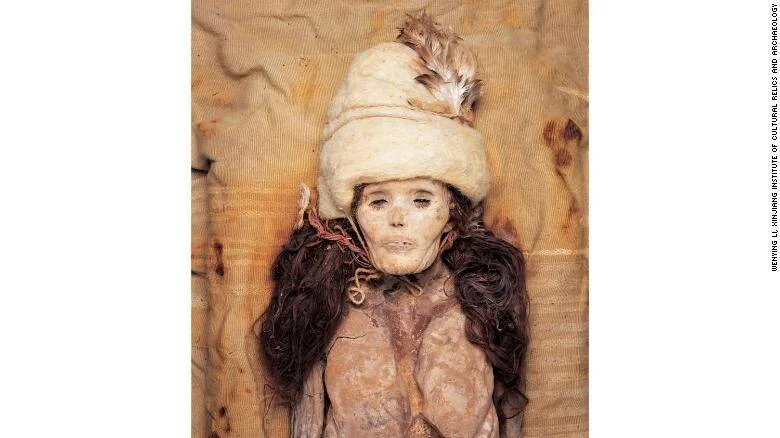 Momia bien preservada con sombrero blanco adornado con plumas y cabello oscuro, hallada en la región de Xinjiang, China.