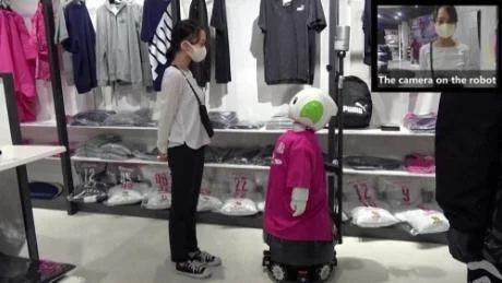 Un robot humanoide interactúa con una clienta en una tienda de ropa Puma, con cámara integrada visible en pantalla secundaria.