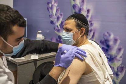 Un hombre con kipá y mascarilla recibe una inyección en el brazo administrada por un sanitario con guantes azules.