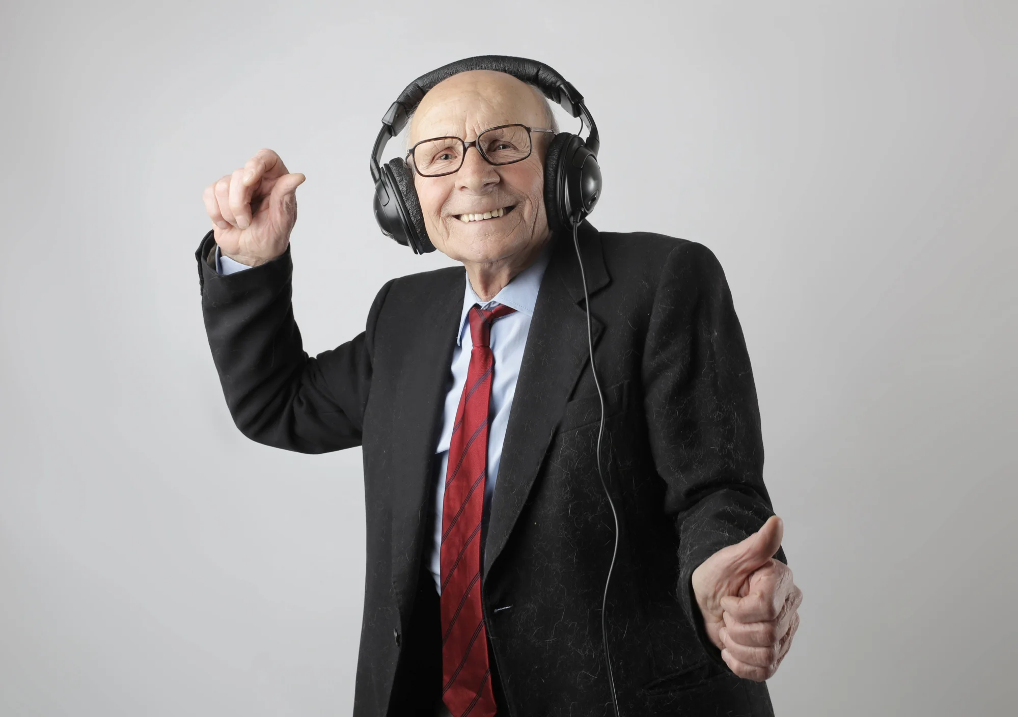 Hombre mayor con traje negro y corbata roja baila sonriente mientras escucha música con auriculares.