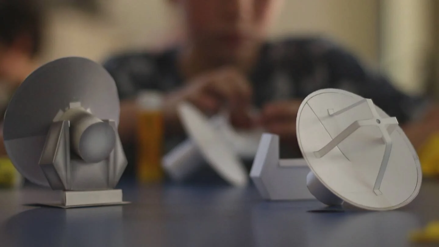 Dos pequeños modelos a escala de antenas parabólicas sobre una mesa, con una persona trabajando al fondo.