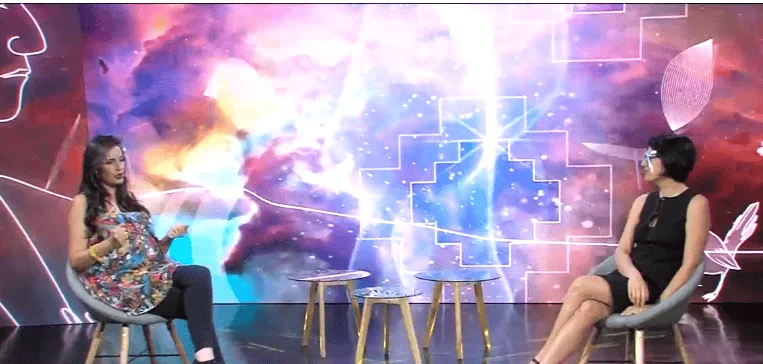 Dos mujeres sentadas frente a frente en un estudio de TV con fondo digital colorido de nebulosa.