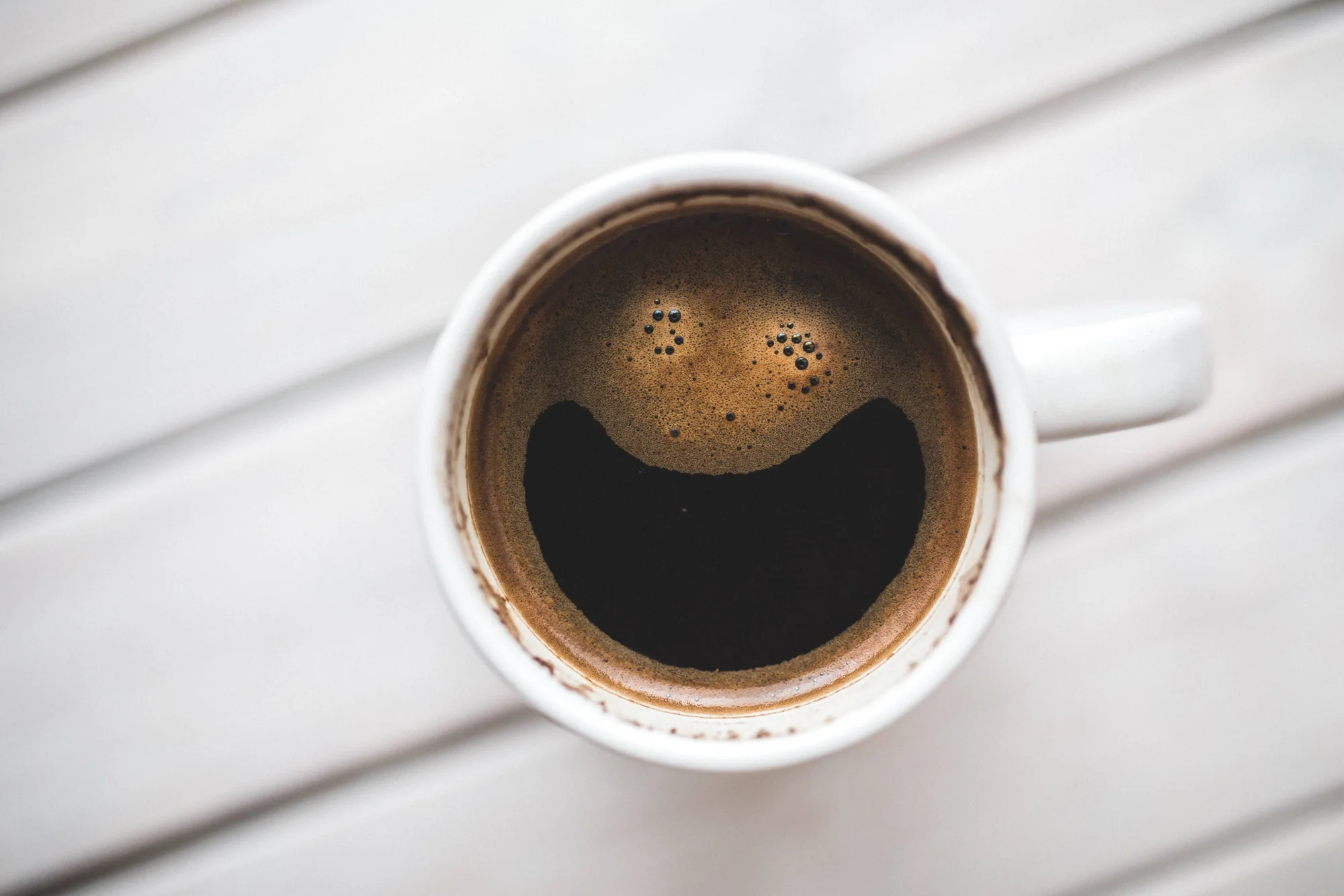 Vista cenital de una taza blanca de café negro cuya espuma forma una cara sonriente.