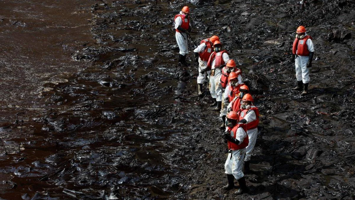 Equipo de trabajadores con trajes blancos y cascos naranja camina sobre una costa cubierta de petróleo durante labores de limpieza.