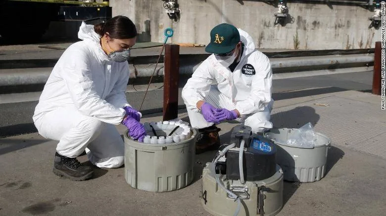 Dos técnicos con trajes de protección y guantes recogen muestras de aguas residuales en contenedores sobre la acera.