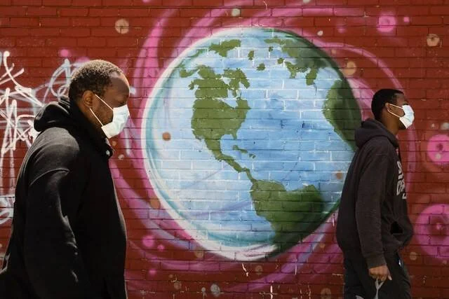 Dos hombres con mascarillas caminan frente a un mural de un globo terráqueo pintado en una pared de ladrillos rojos.