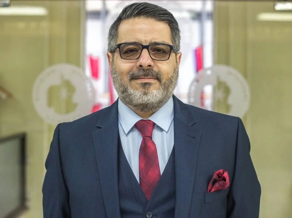 Hombre de mediana edad con barba canosa, traje azul marino, corbata y pañuelo rojo, posando en un entorno institucional.