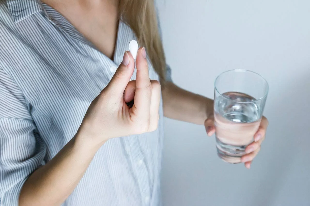 Mujer a punto de tomar una pastilla blanca con un vaso de agua en la mano.