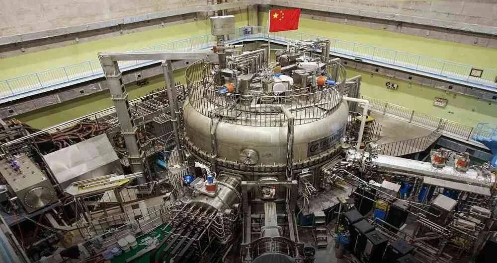 Reactor de fusión nuclear tokamak con bandera china ondeando en su parte superior, rodeado de tuberías y equipos científicos.