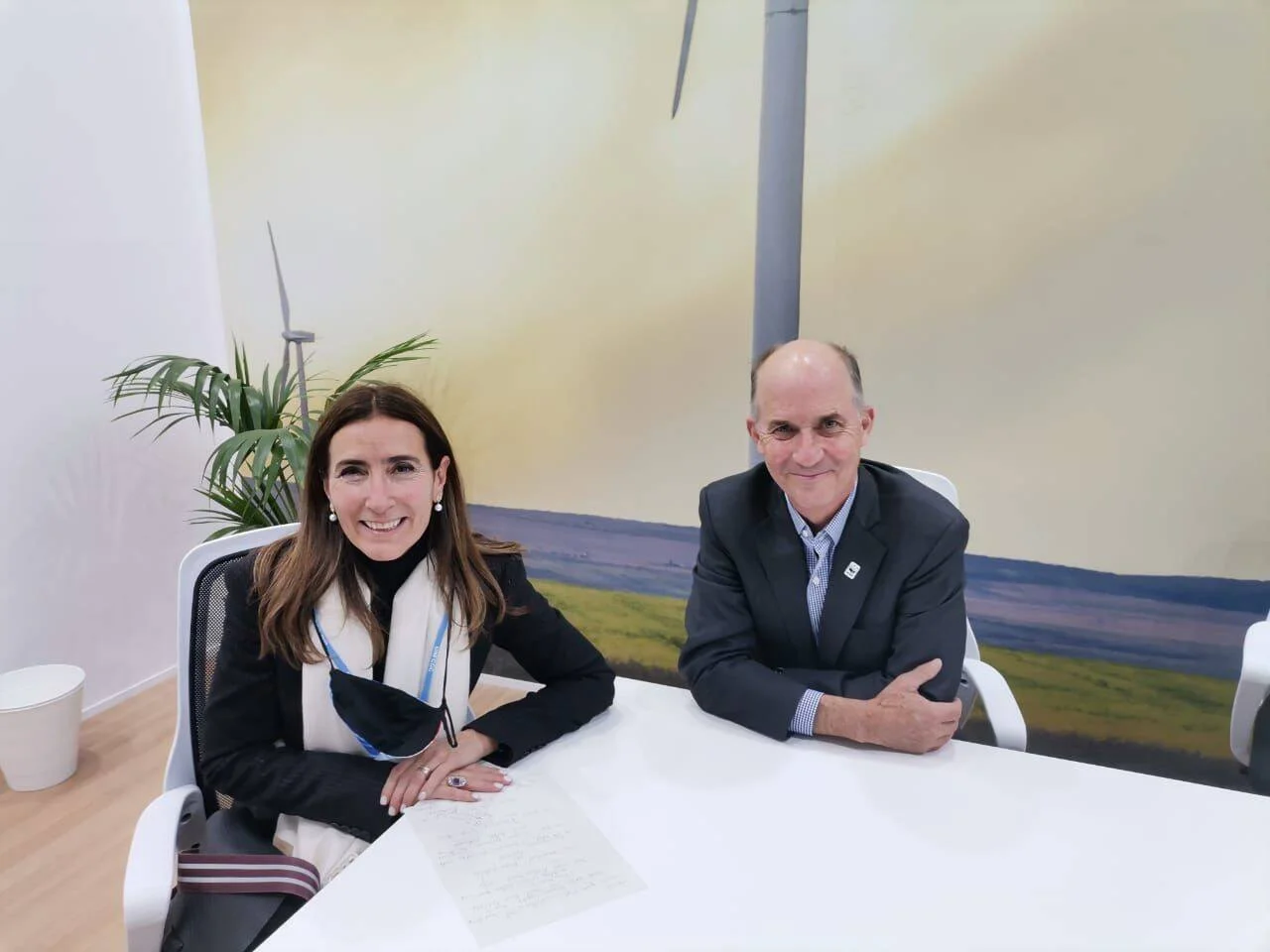 Una mujer y un hombre con credenciales posan sentados ante una mesa blanca, con un mural de aerogeneradores de fondo.