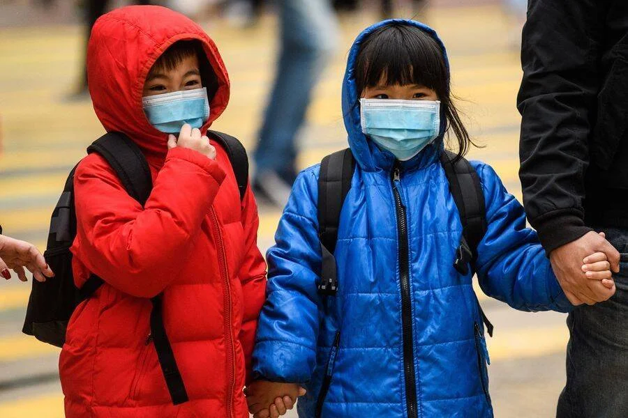 Dos niños pequeños con mascarillas quirúrgicas, abrigos de invierno y mochilas caminan de la mano por una calle.