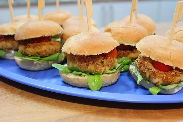 Varios sliders con medallones empanados, tomate y hojas verdes, servidos en una bandeja azul con palillos de madera.
