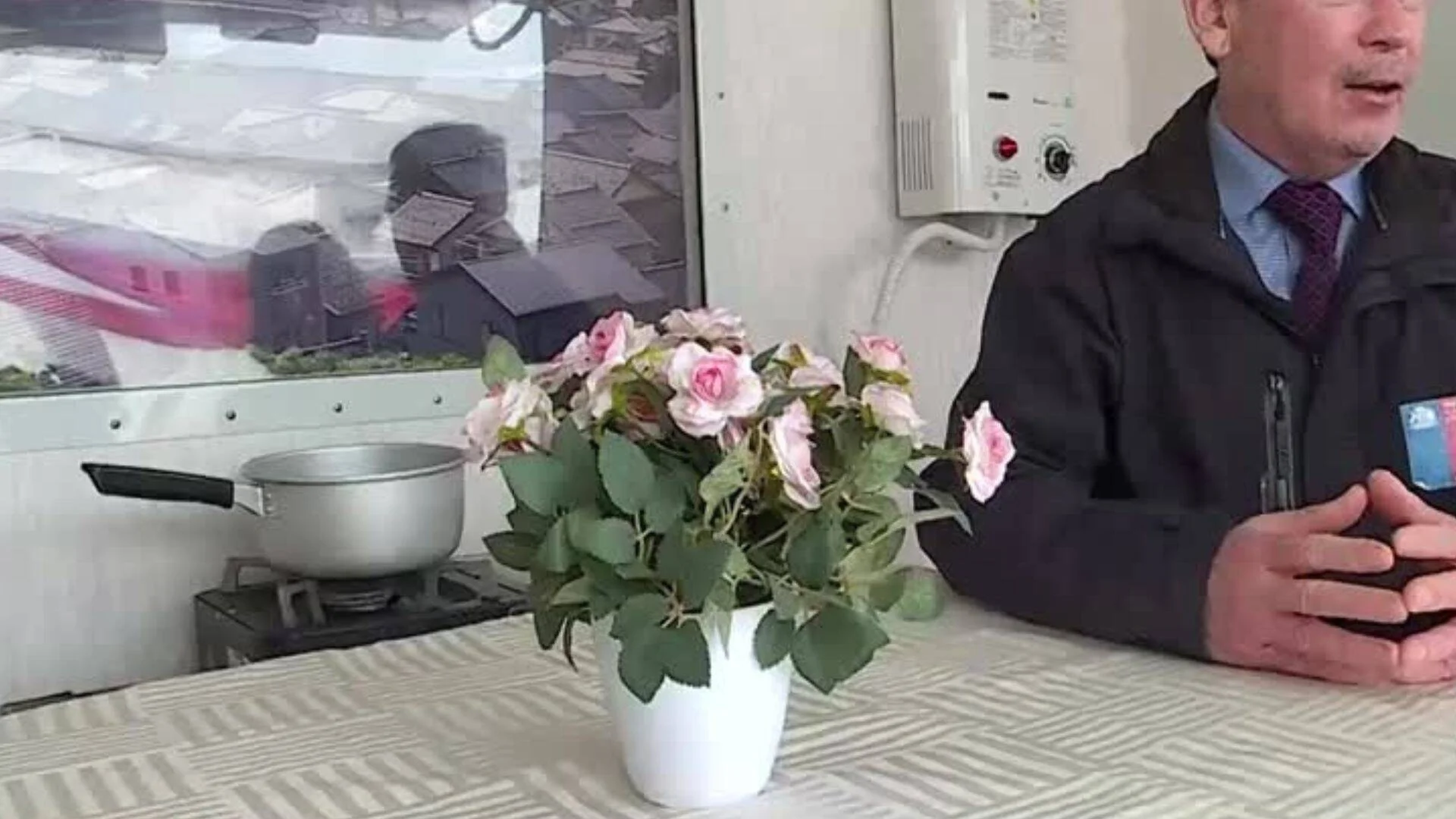 Hombre con traje y chaqueta sentado en una habitación modesta con un ramo de rosas rosadas y una olla sobre una hornalla.