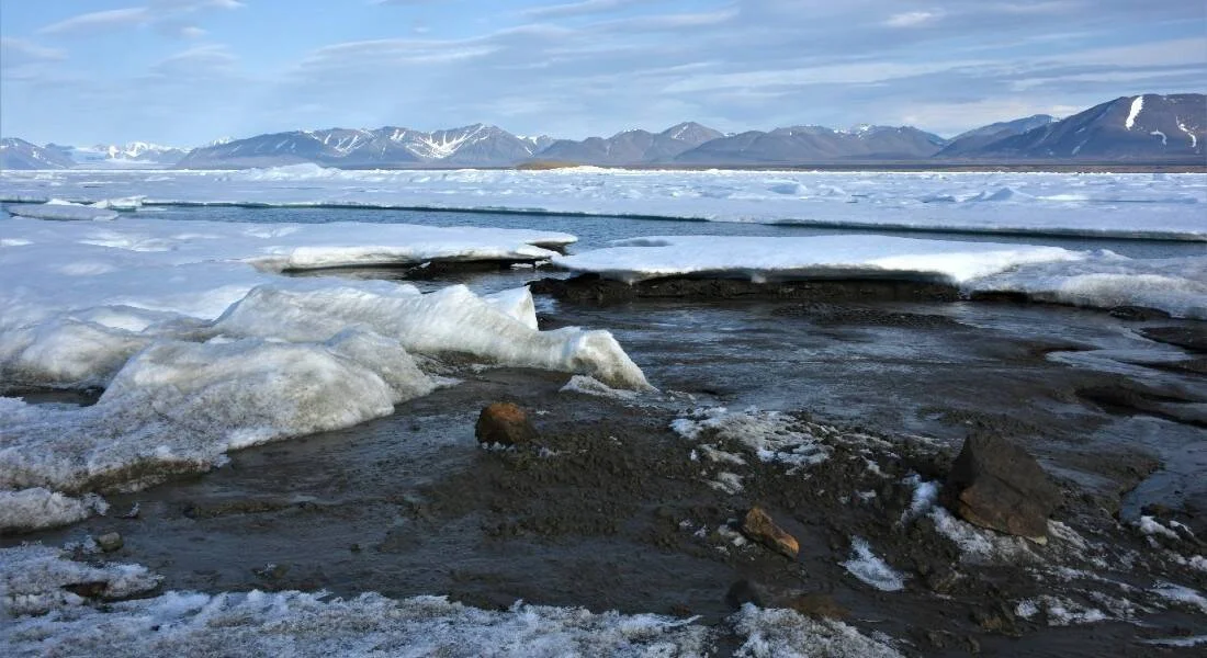 Bloques de hielo derritiéndose sobre una costa rocosa ártica, con placas de hielo marino flotando y montañas nevadas al fondo.