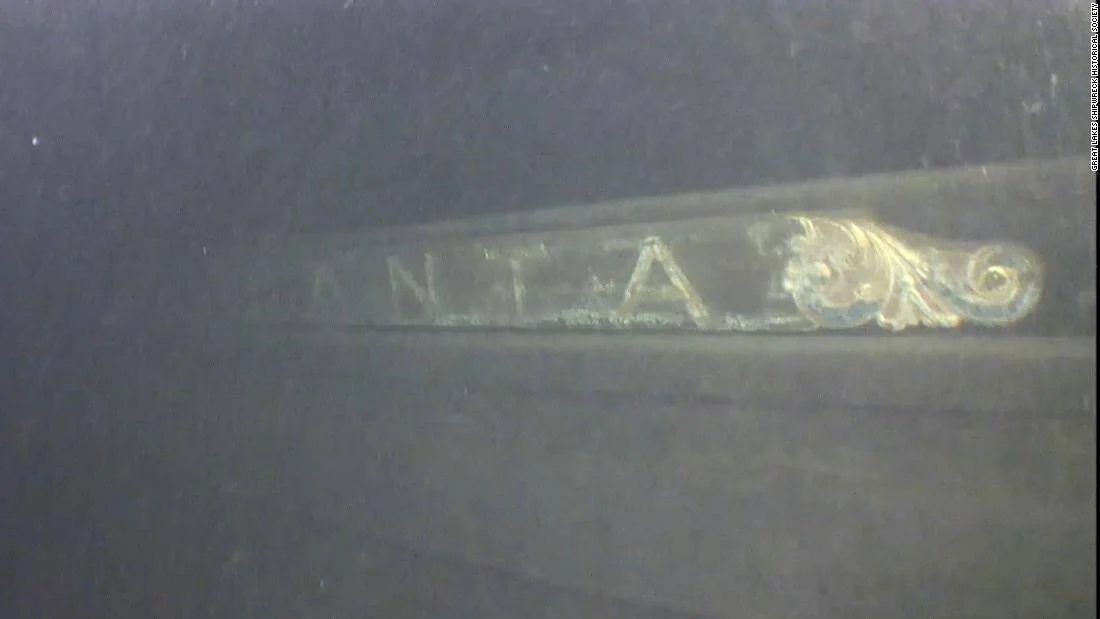 Casco de embarcación hundida fotografiado bajo el agua, con letras parcialmente visibles y ornamento dorado en relieve.