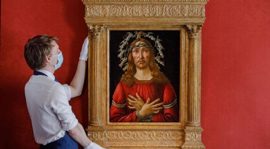 Un técnico con guantes blancos y mascarilla ajusta una pintura renacentista de Cristo con corona de espinas en marco dorado sobre pared roja.