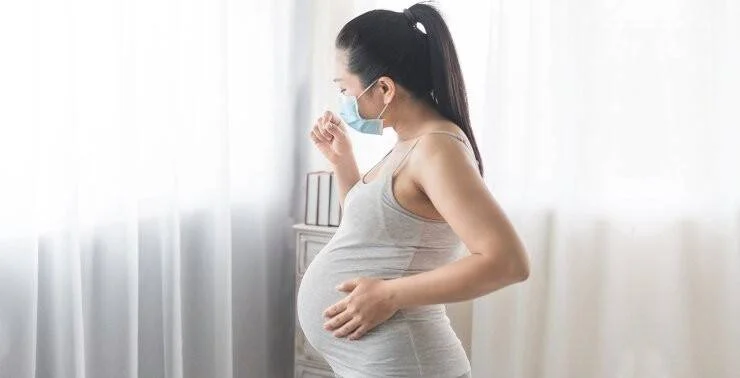 Mujer embarazada con mascarilla quirúrgica tose mientras sostiene su vientre, de perfil junto a una ventana.