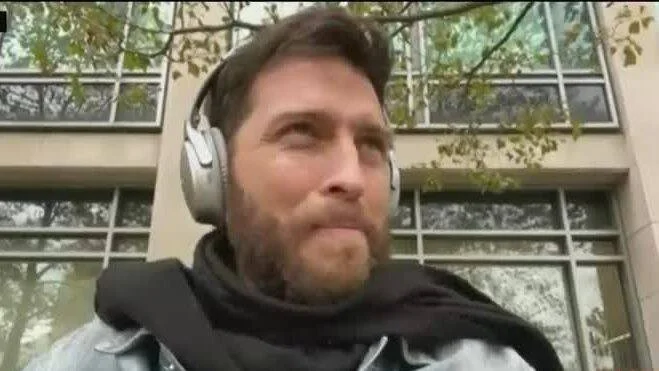 Hombre con barba y auriculares blancos habla frente a un edificio con ventanales.