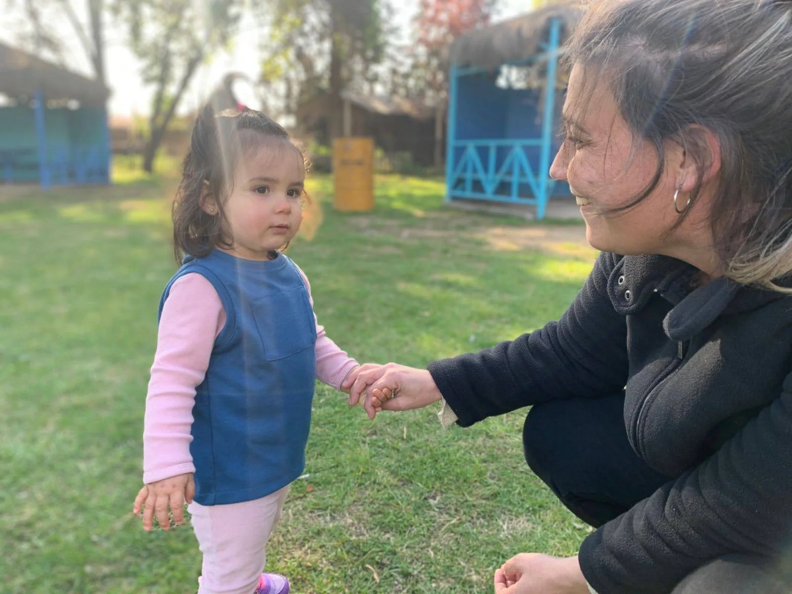 Una mujer adulta se agacha para tomar la mano de una niña pequeña en un parque con césped verde.