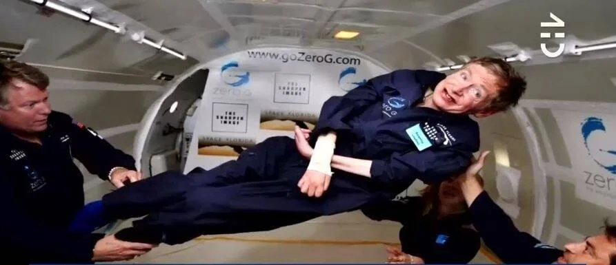 Stephen Hawking flota en ingravidez dentro de un avión de Zero-G, asistido por acompañantes, con una sonrisa en el rostro.