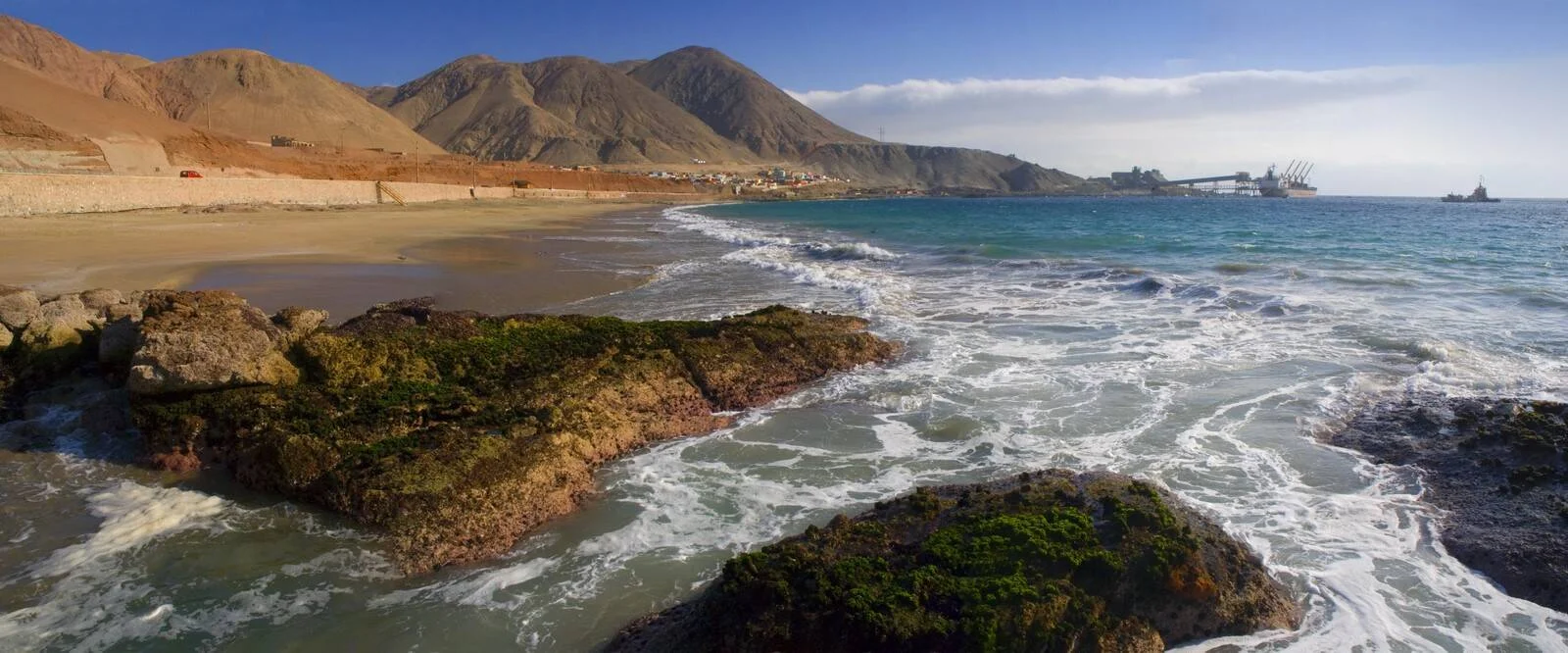 Playa rocosa con oleaje frente a montañas áridas y una instalación portuaria al fondo en una bahía de aguas turquesas.