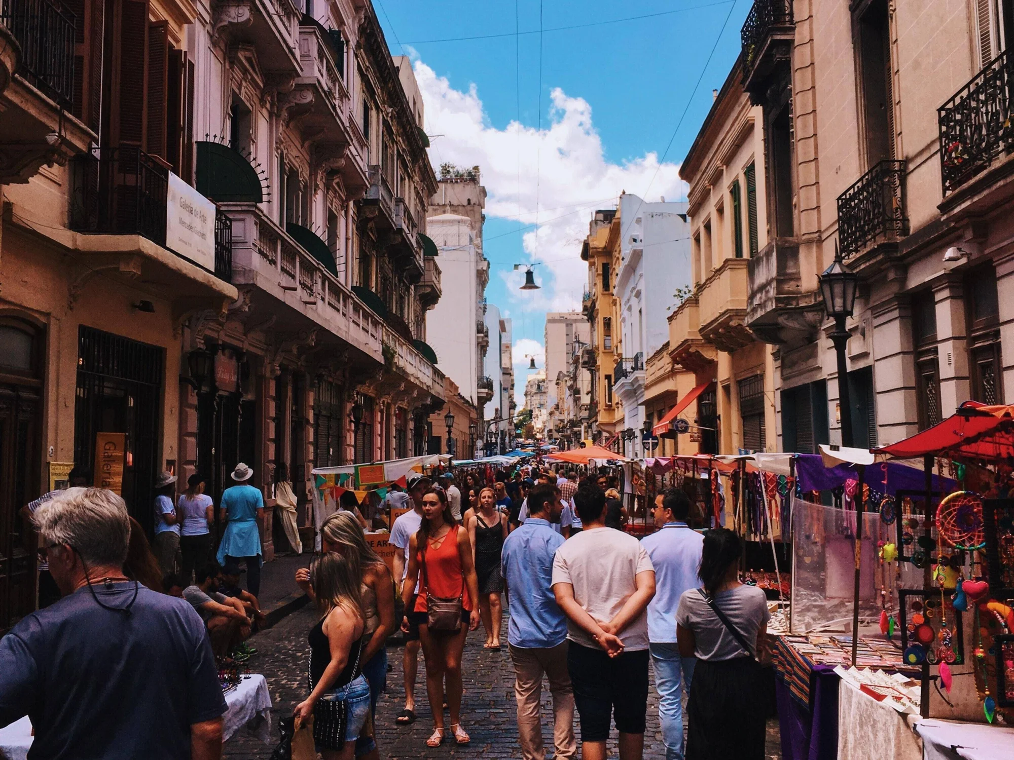 Multitud de personas recorre una feria artesanal en una calle adoquinada flanqueada por edificios coloniales bajo cielo azul.