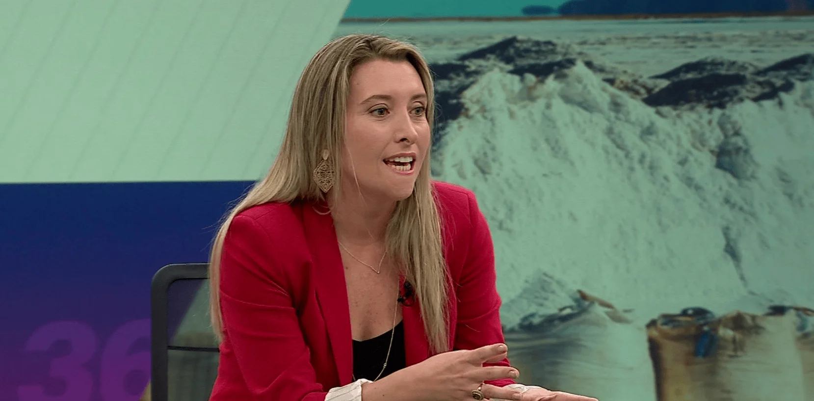 Mujer rubia con saco rojo habla en un estudio de televisión con imagen de salinas al fondo.