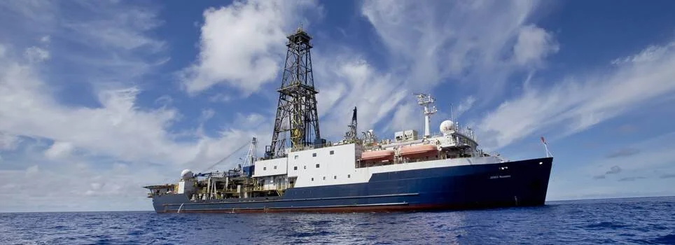 Buque de perforación científica con torre de perforación navegando en aguas oceánicas bajo cielo parcialmente nublado.