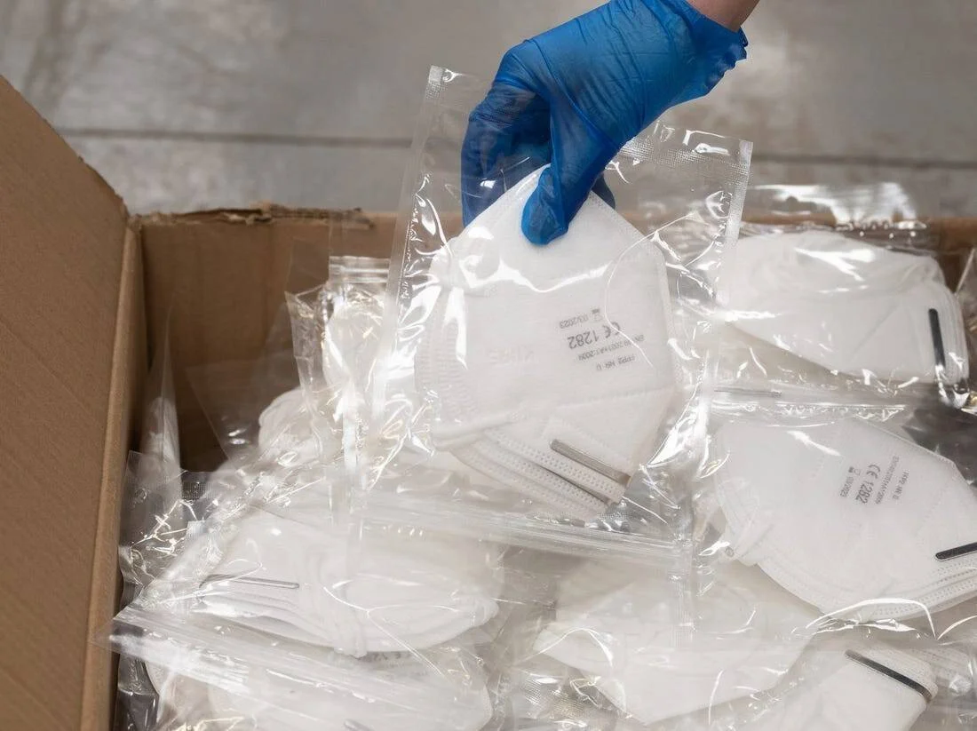 Una mano con guante azul extrae una mascarilla FFP2 sellada individualmente de una caja de cartón llena de ellas.