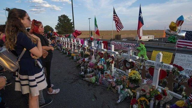 Personas observan un memorial con cruces blancas, flores y banderas de México y Texas en honor a las víctimas de un tiroteo masivo.