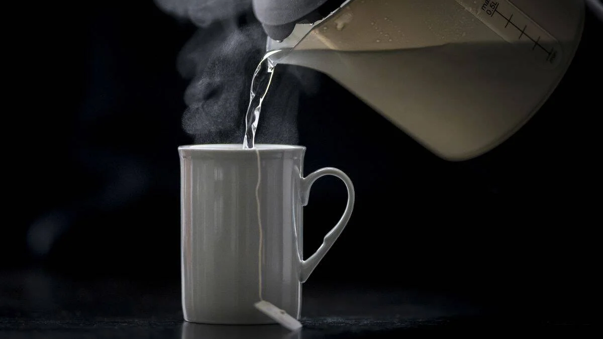 Agua caliente siendo vertida desde una jarra medidora en una taza blanca, con vapor visible sobre fondo oscuro.