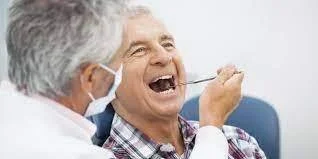 Dentista con mascarilla examina la boca abierta de un hombre mayor con espejo dental.