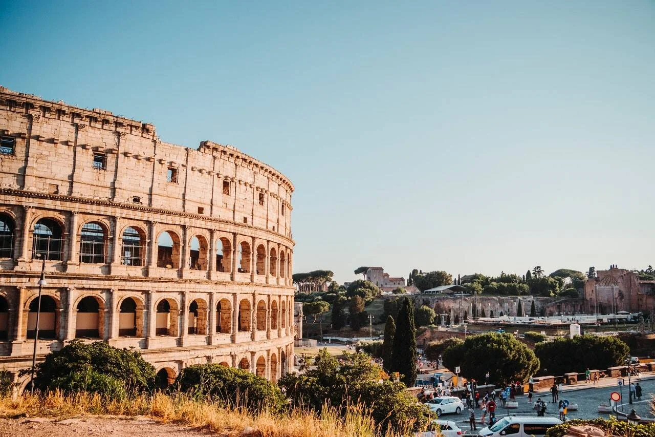 Vista exterior del Coliseo de Roma con turistas y el monte Palatino al fondo bajo un cielo azul despejado.