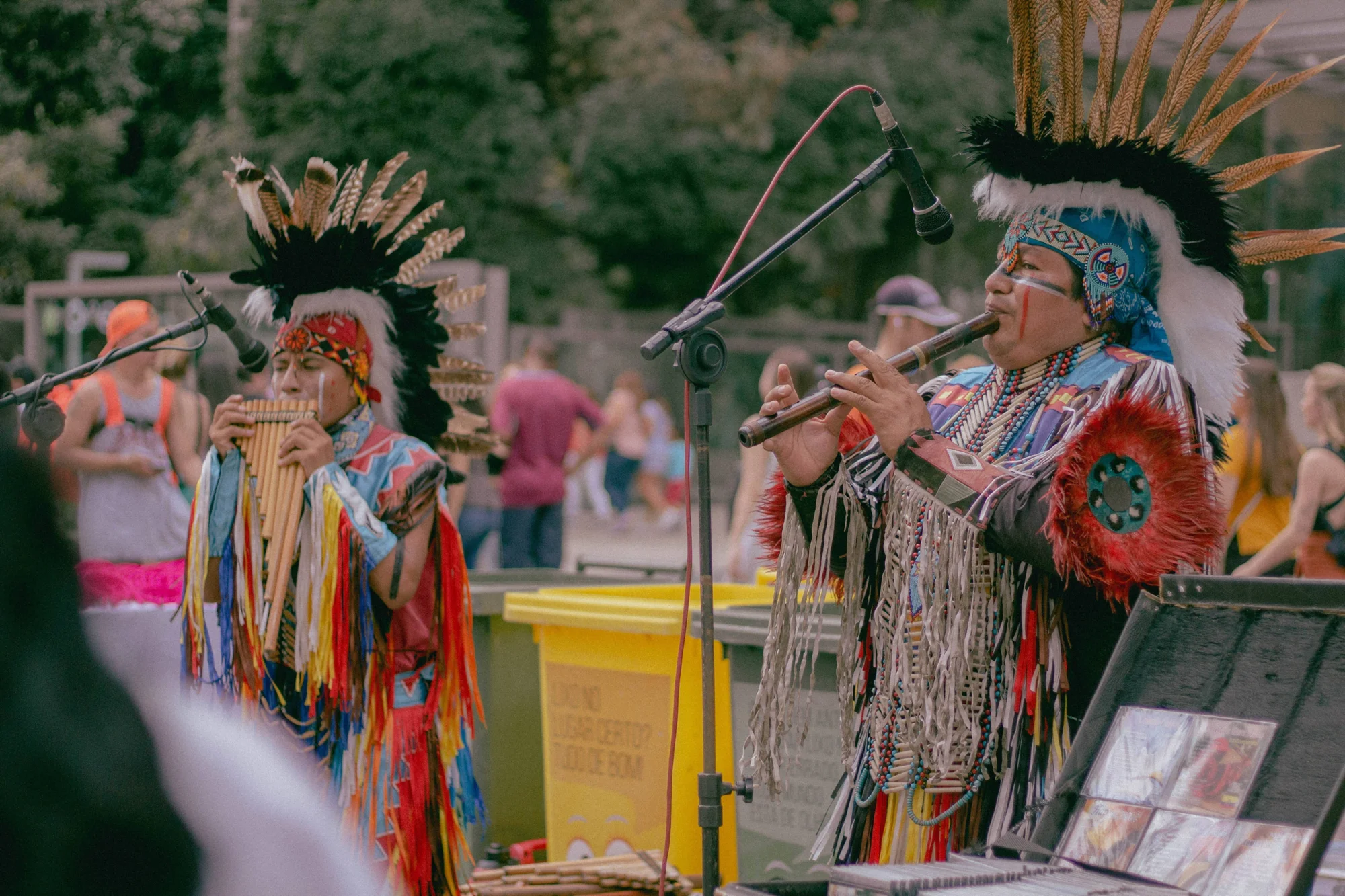 Dos músicos con atuendos y tocados de plumas indígenas tocan zampoña y flauta ante micrófonos en un espacio público.