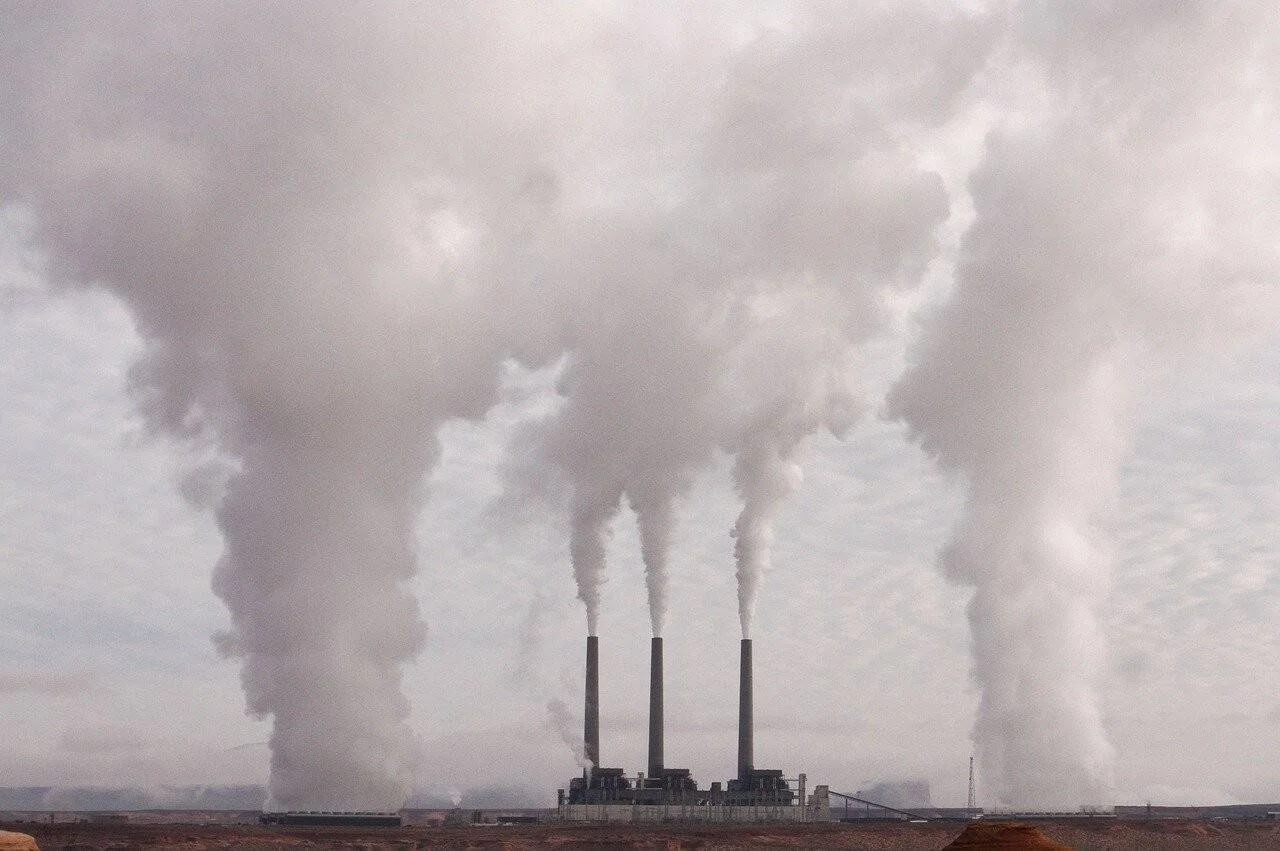 Tres chimeneas de una planta industrial expulsan grandes columnas de humo gris que cubren el cielo en un paisaje árido.