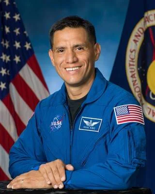 Astronauta con traje azul de la NASA posa en retrato oficial frente a banderas estadounidense y de la agencia espacial.
