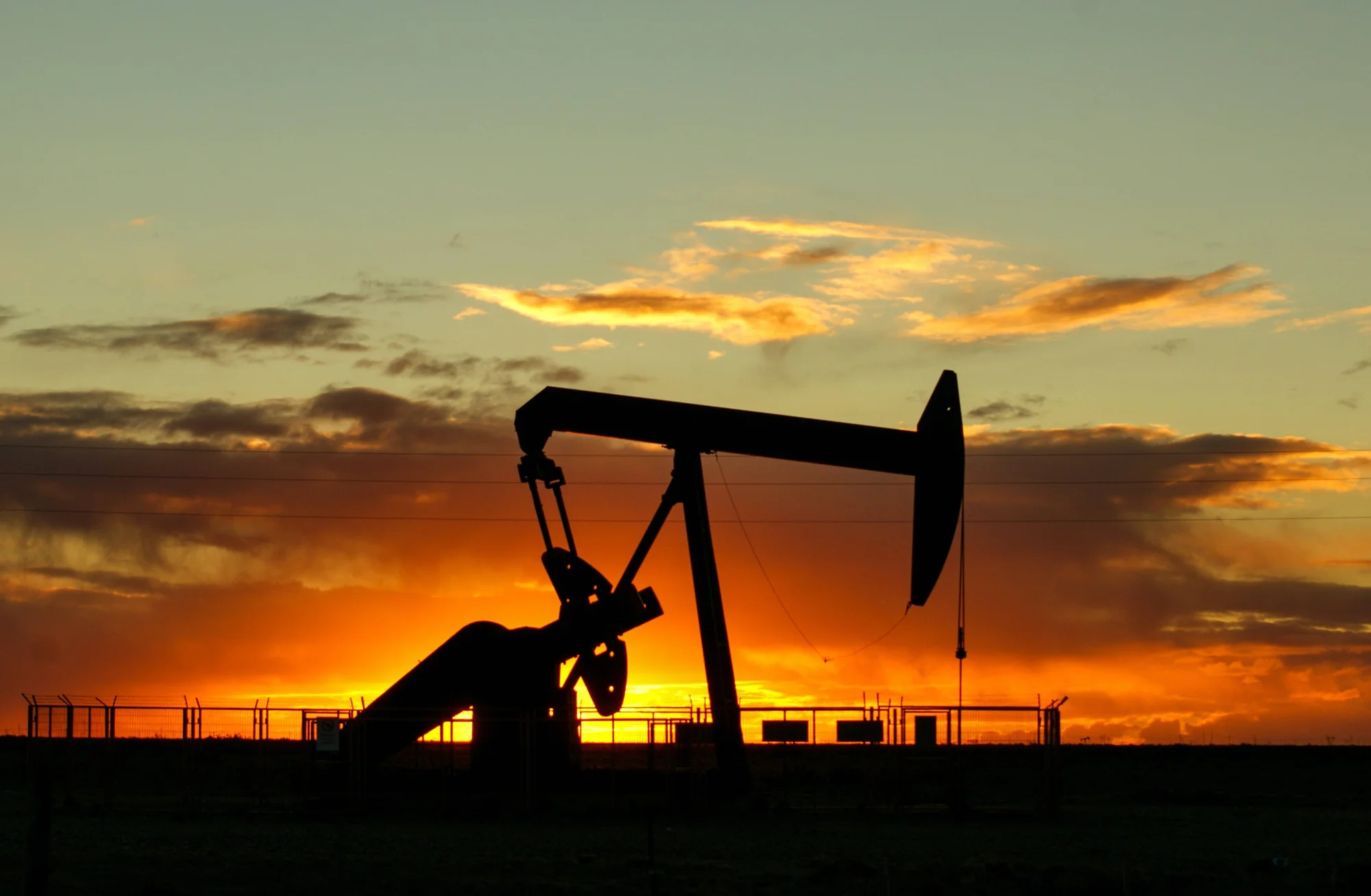 Silueta de una bomba extractora de petróleo contra un cielo naranja al atardecer, con instalaciones industriales al fondo.
