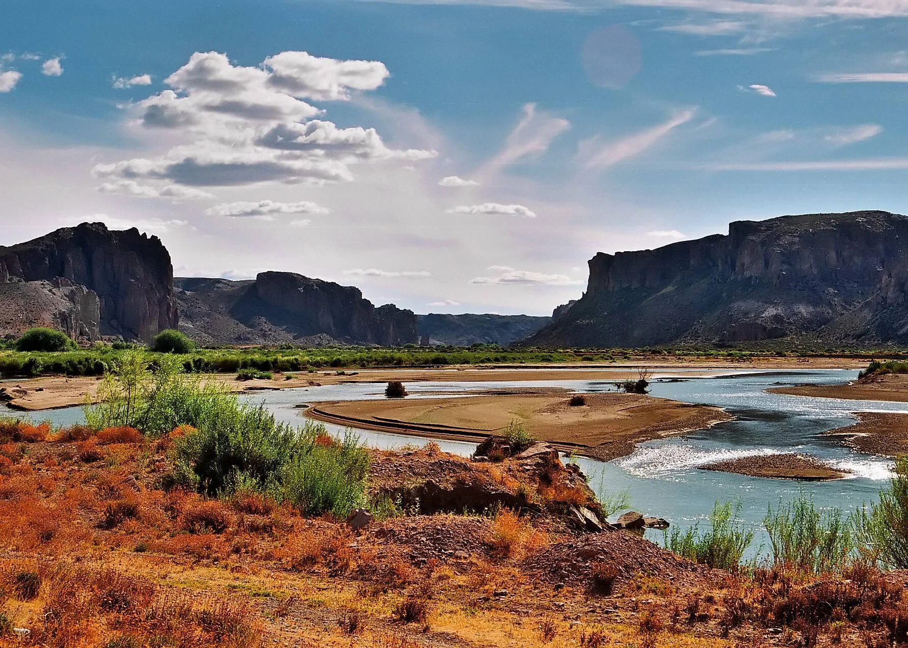 Río de aguas turquesas serpentea entre acantilados rocosos y vegetación árida bajo un cielo parcialmente nublado.
