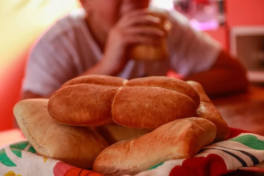 Varios panes de flautita recién horneados apilados sobre un mantel floreado, con una persona comiendo al fondo.