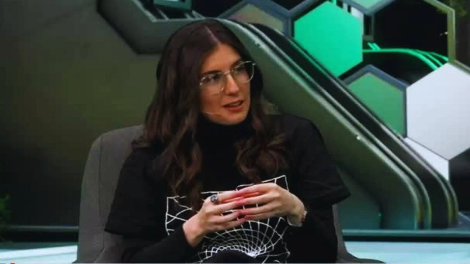 Mujer joven con cabello oscuro, anteojos y remera negra habla durante una entrevista en un estudio de televisión con decorado verde.
