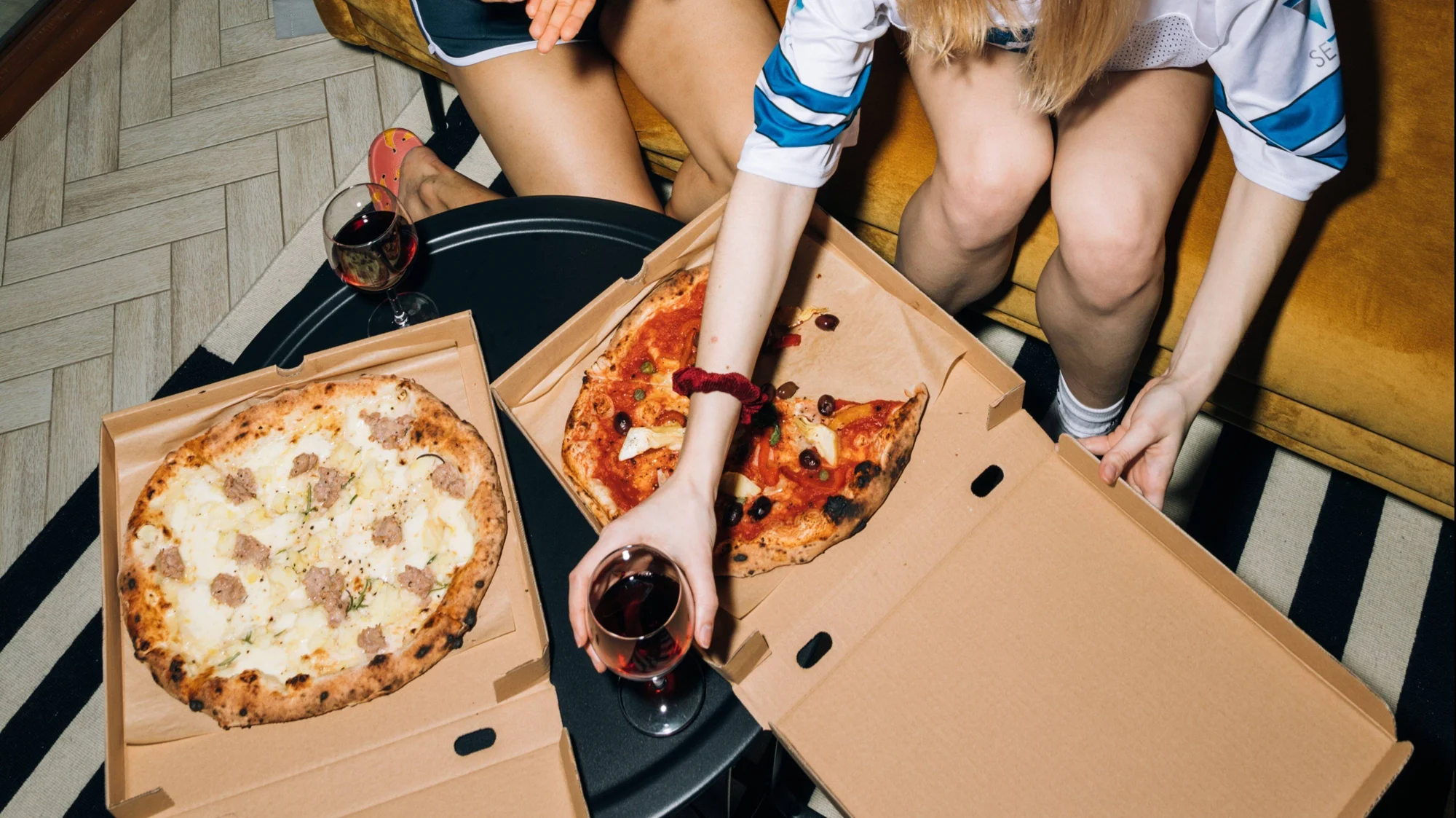 Vista cenital de dos pizzas artesanales en cajas de cartón sobre mesa, acompañadas de copas de vino tinto y dos personas sentadas.